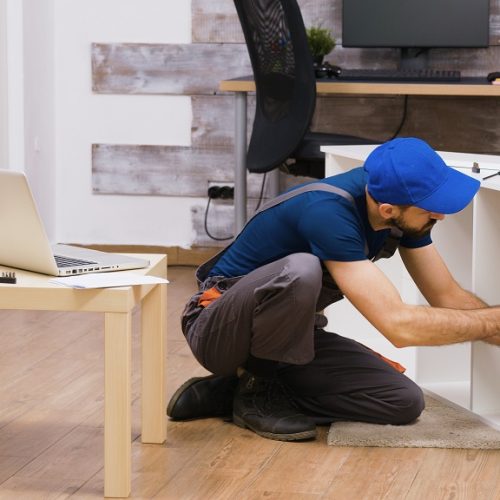 Professional worker in overalls consults furniture assembly instructions from laptop. Worker wearing a cap.